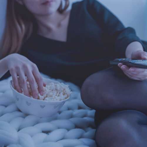 woman in white bed holding remote control while eating popcorn